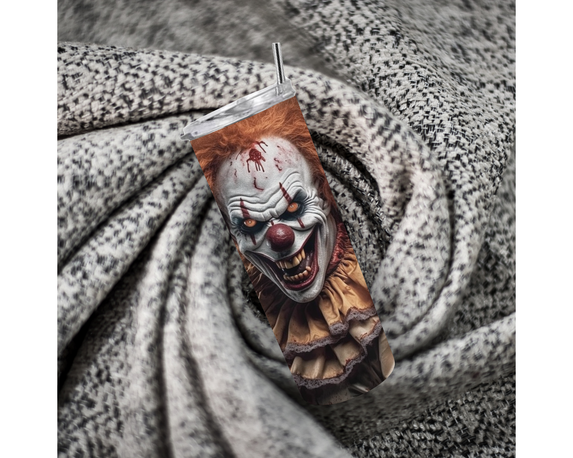 A skinny tumbler with a printed horror clown design, featuring a metal straw, resting on a textured gray and white patterned fabric background.