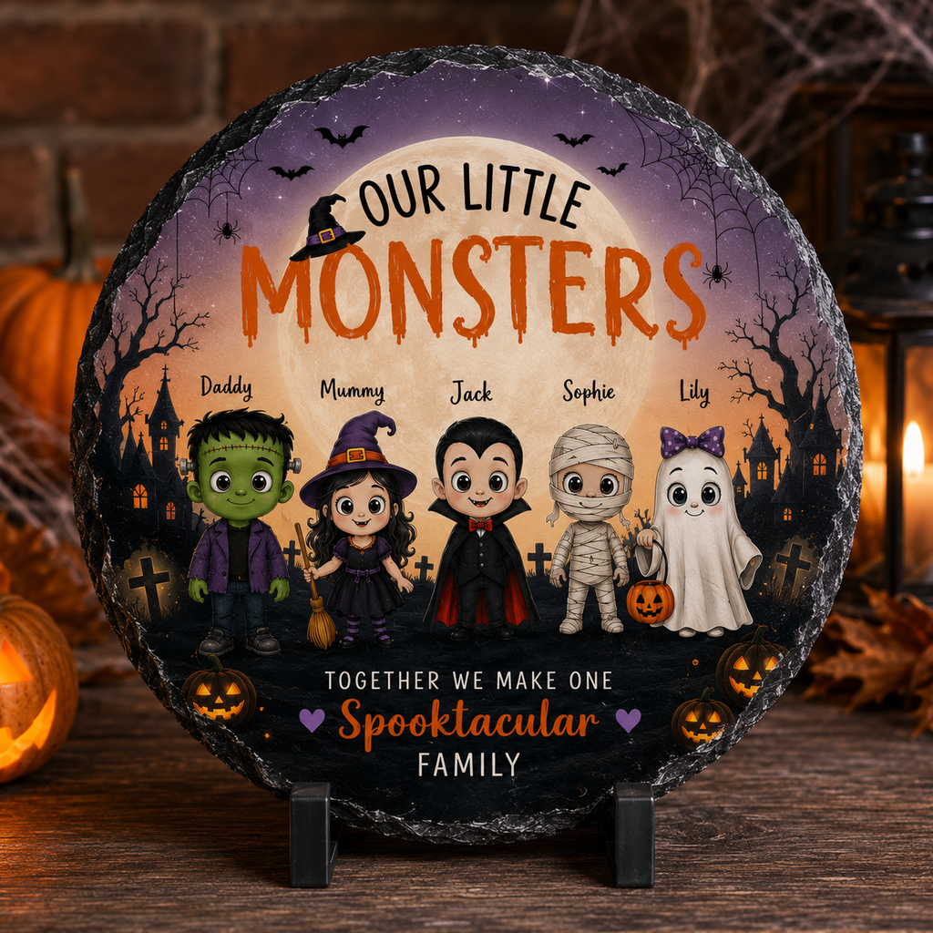 A round personalized slate plaque with a Halloween design, displayed on a small stand on a wooden surface with a cozy, warm background of candles, lanterns, and a brick wall. The plaque features cartoon characters resembling Frankenstein's monster, a witch, vampire, mummy, and ghost, with customizable names and the text 'OUR LITTLE MONSTERS' and 'TOGETHER WE MAKE ONE Spooktacular FAMILY'.