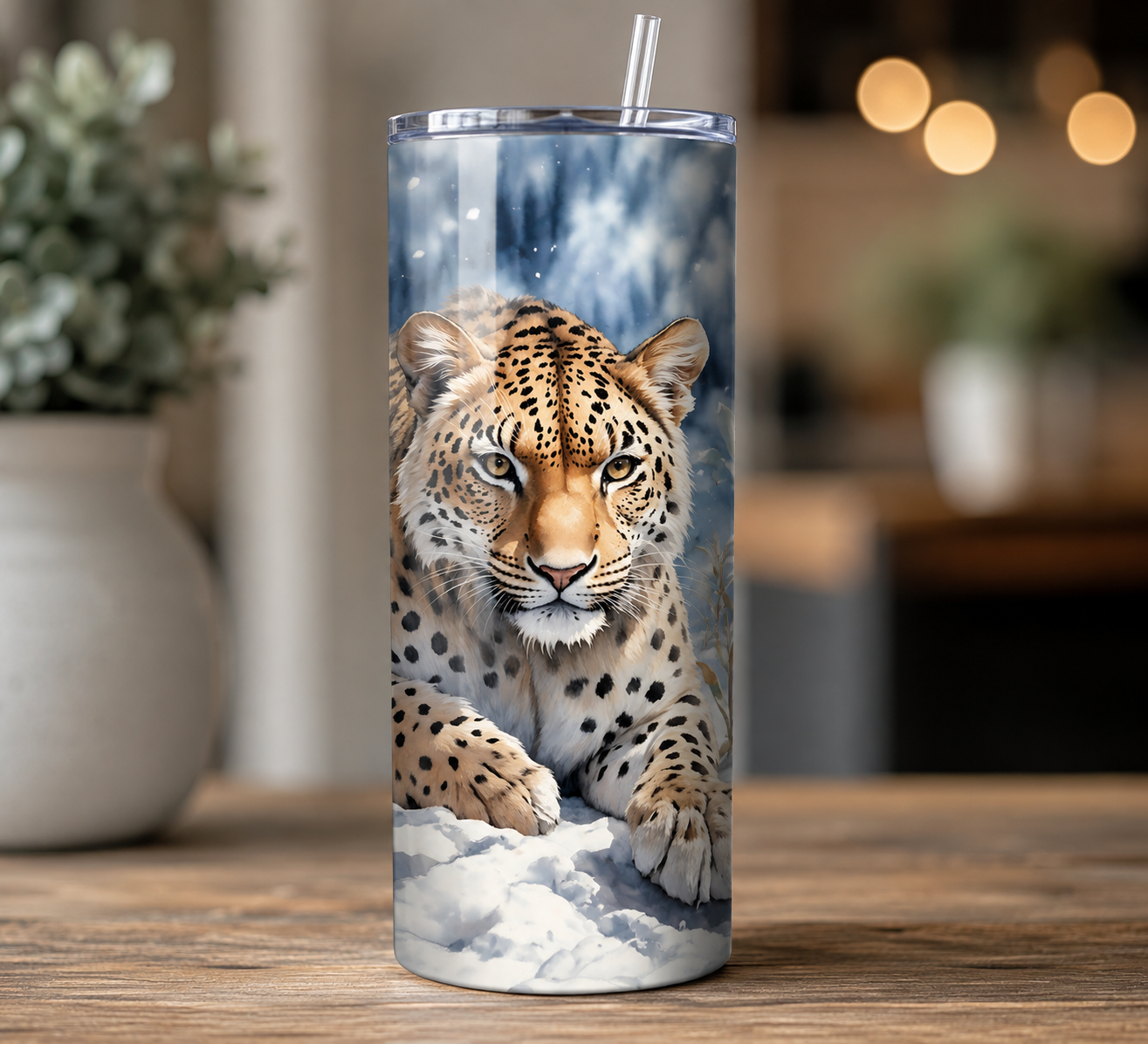 A tall insulated tumbler with a clear lid and straw, featuring a printed leopard over a snowy blue-and-white background, shown on a wooden table in a softly blurred indoor setting.