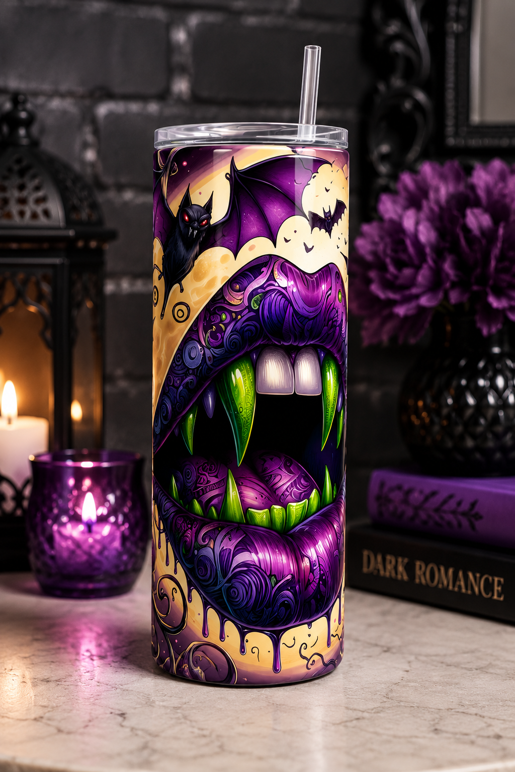 A tall insulated tumbler with a clear lid and straw, featuring a Halloween-themed graphic of an open mouth with fangs, purple lips, bats, and ghosts. The tumbler is displayed on a marble surface with gothic decor props including a candle, lantern, and books in the background.