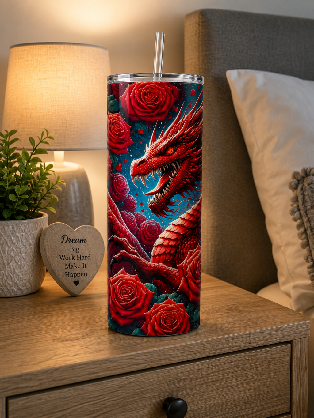 A tall insulated tumbler with a clear lid and straw sits on a wooden bedside table in a bedroom setting. The tumbler features a vivid printed design of a red dragon surrounded by red roses on a dark blue background.