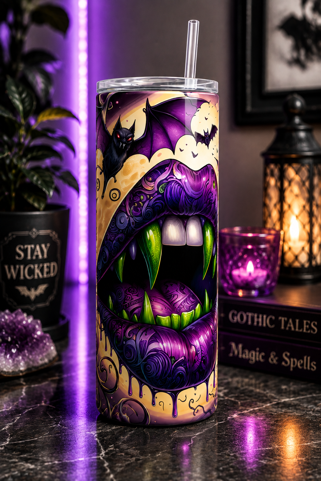 A tall insulated tumbler with a clear lid and straw, featuring a vivid Halloween-style monster mouth illustration in purple, black, cream, and green. The tumbler is photographed on a dark tabletop with gothic decor props including a plant in a black pot reading 'STAY WICKED,' a lantern, a lit candle, and books.