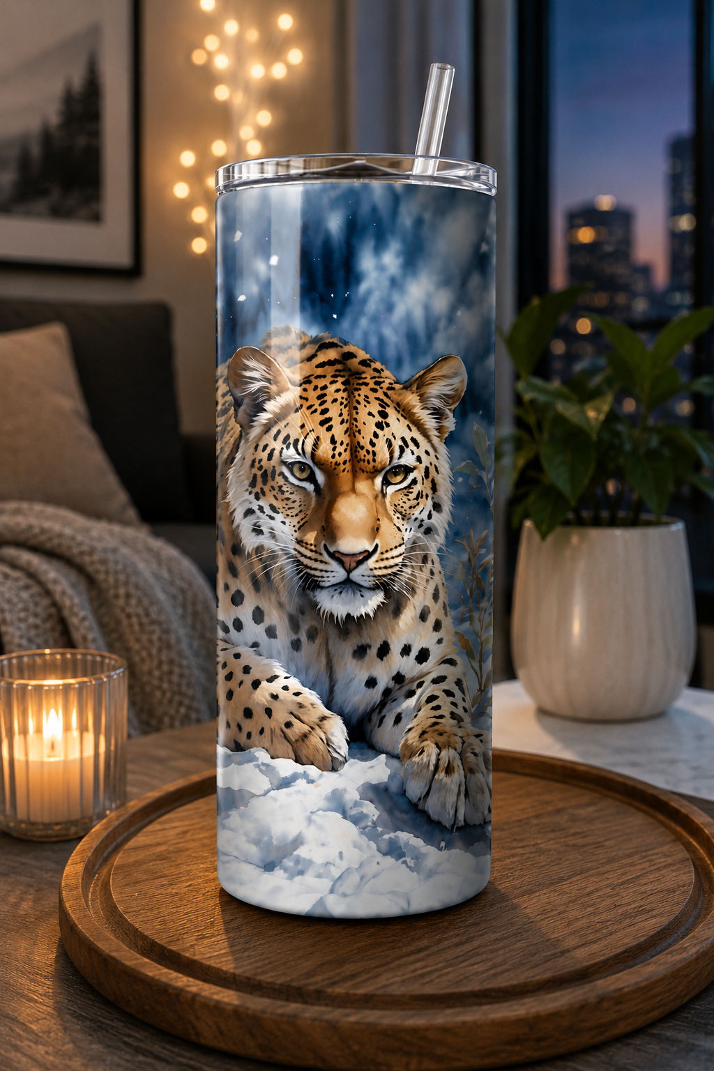 A tall insulated tumbler with a clear lid and straw, printed with a realistic leopard over snowy ground, shown standing on a wooden tray in a warmly lit living room.