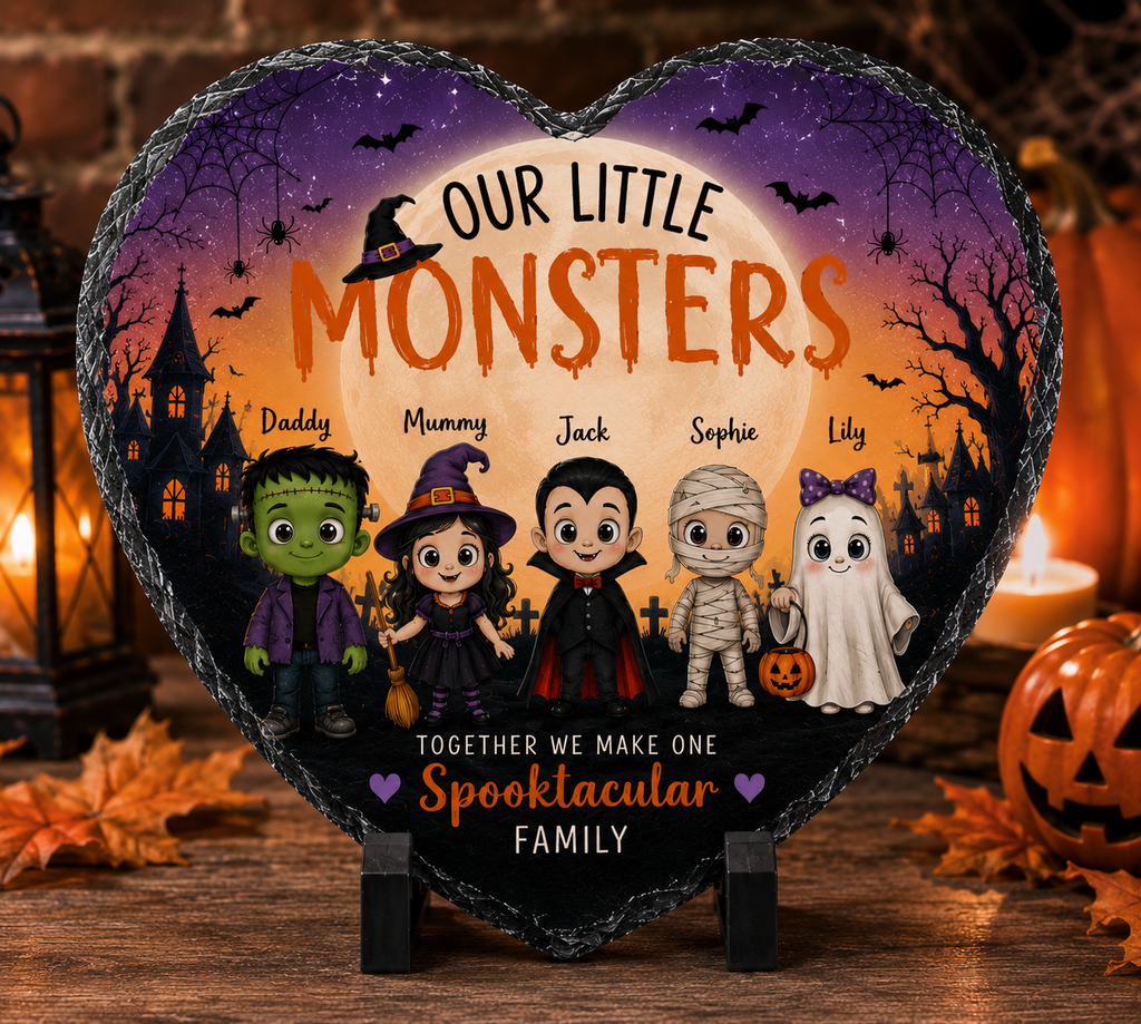 A heart-shaped slate plaque on a small stand, printed with a Halloween family scene and the text 'OUR LITTLE MONSTERS' and 'TOGETHER WE MAKE ONE Spooktacular FAMILY,' shown in a styled holiday setting with candles, lanterns, and pumpkins.