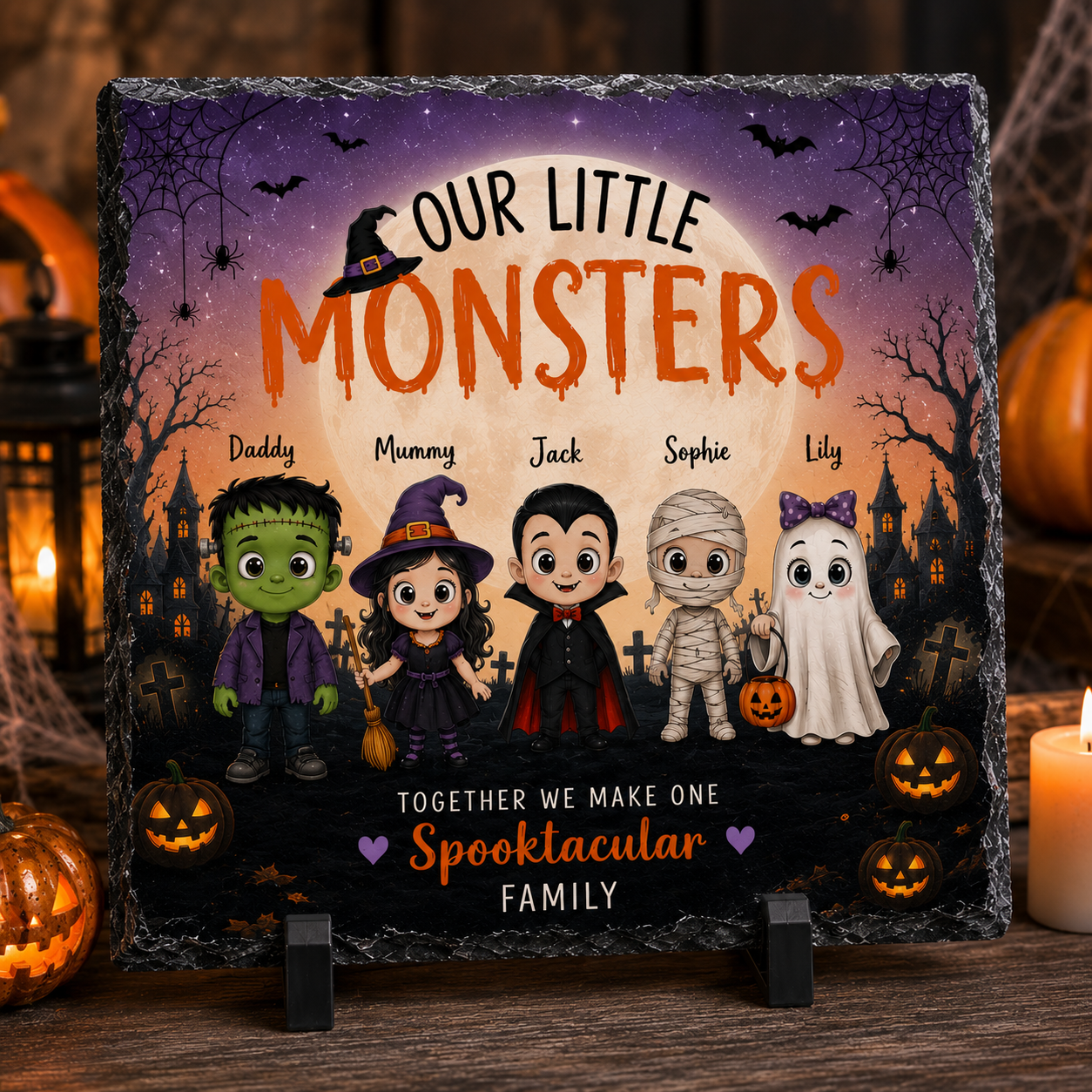 A personalized Halloween-themed square slate plaque on a small black stand, displayed on a wooden surface with candles and pumpkins in the background. The plaque features cartoon characters of a Frankenstein monster, a witch, vampire, mummy, and ghost under a full moon, with customizable text reading 'OUR LITTLE MONSTERS' and 'TOGETHER WE MAKE ONE Spooktacular FAMILY'.
